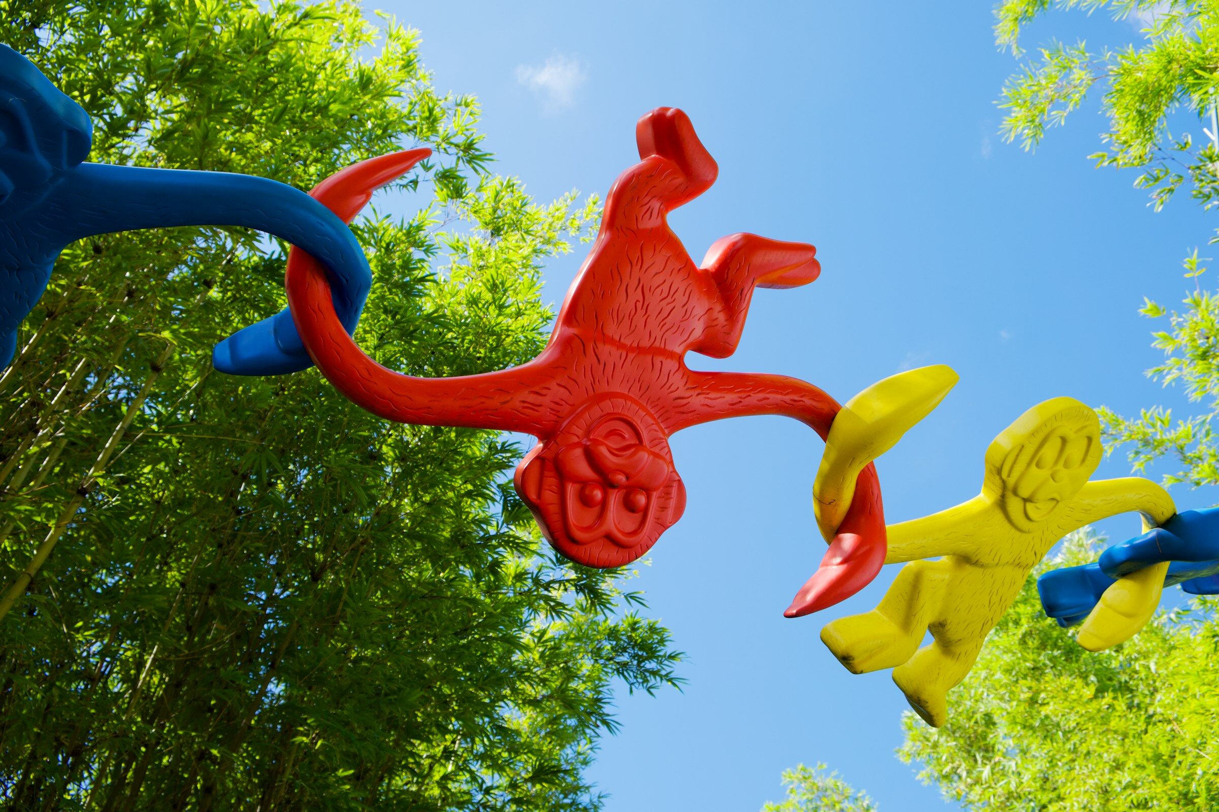 Affen-Figuren aus Plastik in verschiedenen Farben, die ineinander greifen. Im Hintergrund sind Bäume und Himmel zu sehen.
