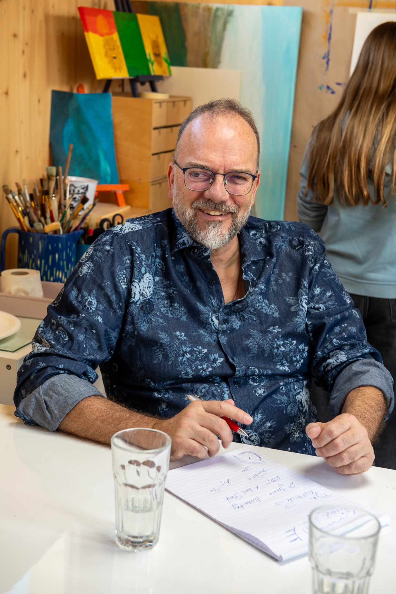 Urs-Micheal Hoenner sitzt lachend im Schulzimmer mit Kinder und macht sich Notizen