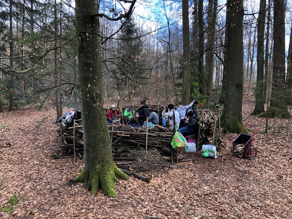 Ein Waldsofa im Wald, gebaut aus Ästen. Die Schülern Schüler sitzen auf dem Waldsofa im Kreis